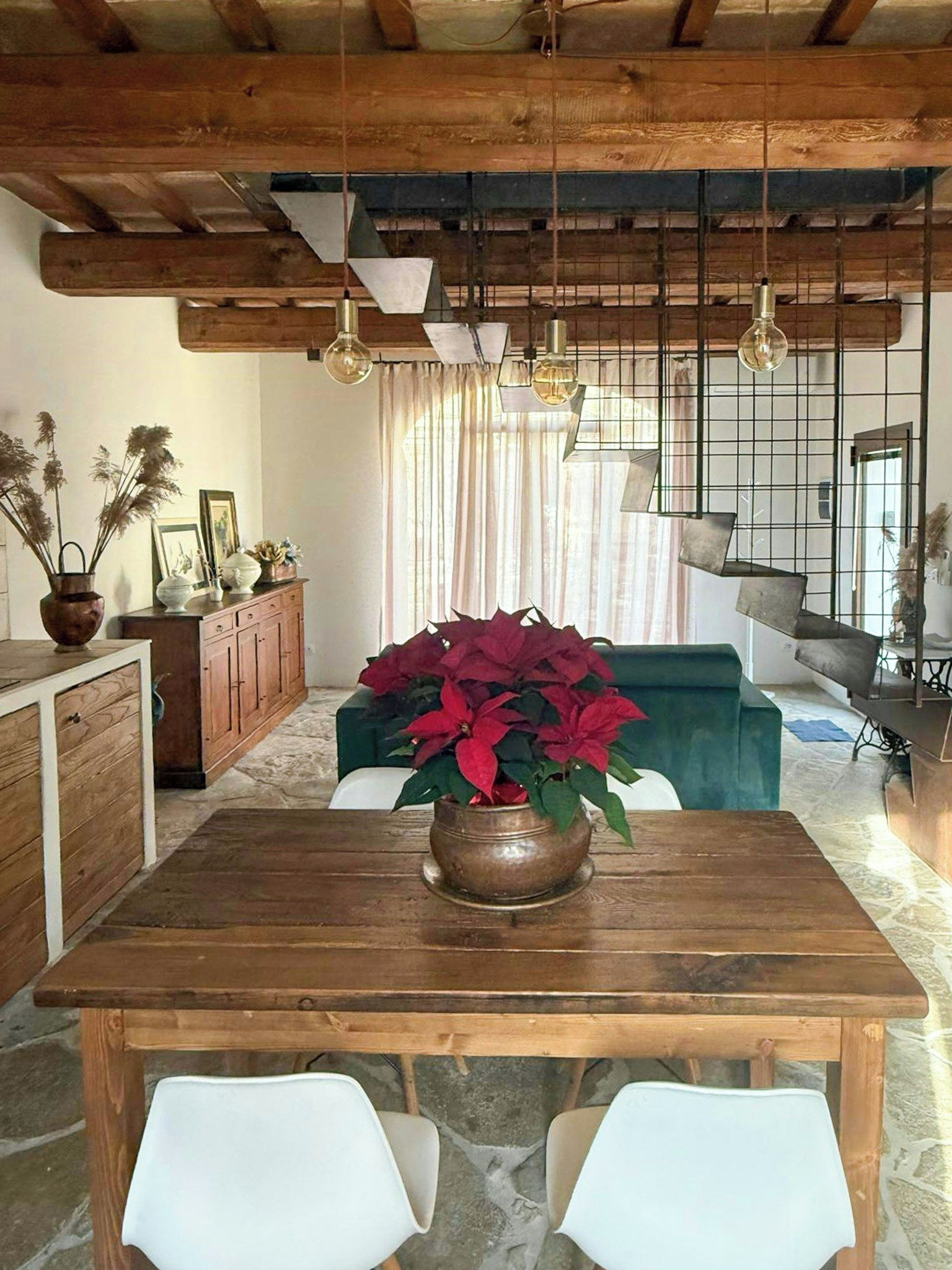 Une table à manger en bois, agrémentée d’une plante en pot, des chaises blanches, un canapé vert et un coin cuisine avec des meubles en bois et un luminaire suspendu.