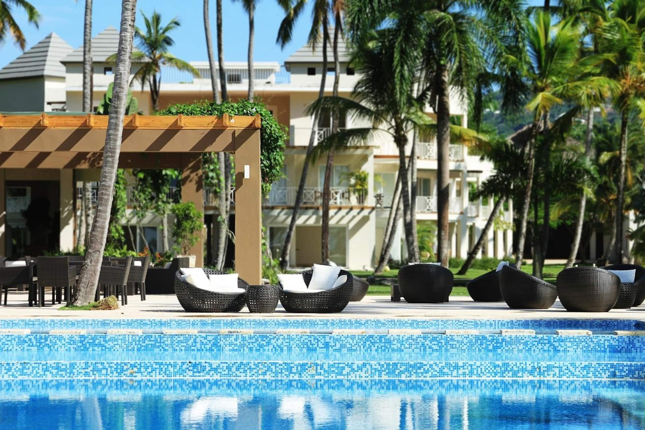 Zona de piscina con sillones reclinables y un área de estar cubierta, rodeada de palmeras y un edificio al fondo.