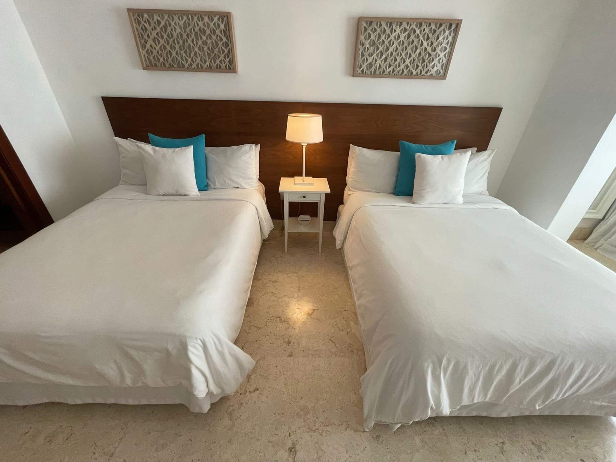 Habitación de hotel con dos camas, una mesita de noche con lámpara y paneles decorativos en la pared.