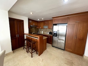 A kitchen with wooden cabinets, a stainless steel refrigerator, a granite countertop, and two bar stools. - Case Damare Coson bay (Las Terrenas)