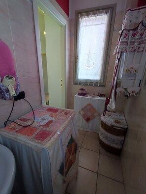 A small, compact room with a table covered in a floral tablecloth, a window with a decorative curtain, and a wall-mounted heater. - MAISON PLATONE CENTER (Alessandria)