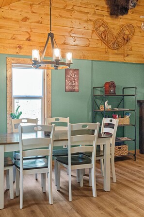 Ein Interieur einer Holzhütte mit Holzbalkendecke, einem Kronleuchter, einem Esstisch mit Stühlen, einer grünen Wand und einem Regal mit verschiedenen Gegenständen.