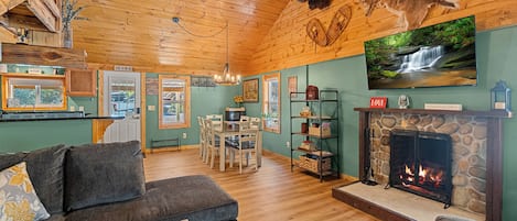 A cozy cabin interior with a fireplace, wooden floors, a dining area, and a television mounted on the wall.