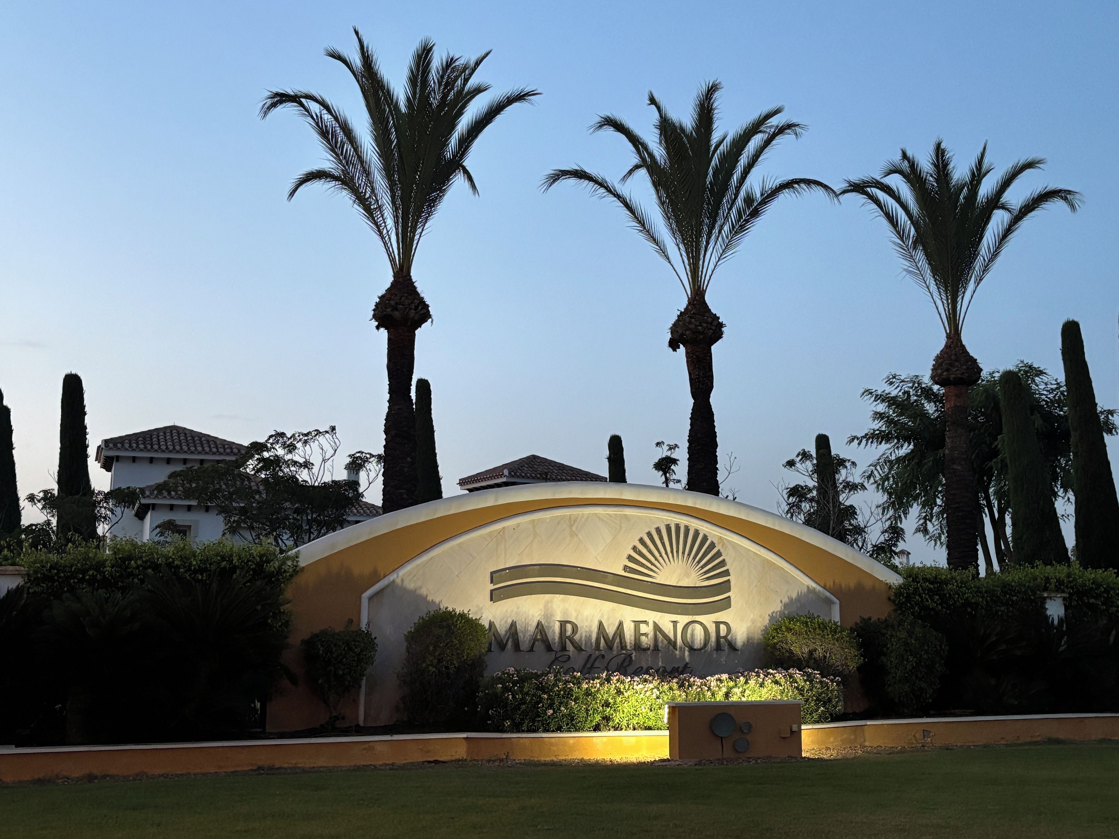 A sign for Mar Menor Golf Resort with palm trees and a building in the background.