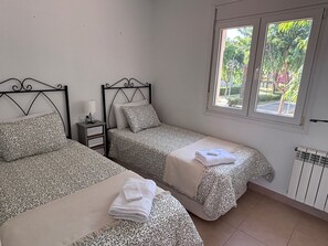 A room with two beds, each with patterned bedding and pillows, a nightstand with a lamp, and a window with a view of greenery.