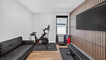 A modern room with a black leather sofa, an exercise bike, a wall-mounted TV, and a wooden panel wall.