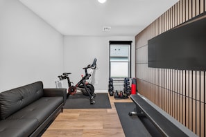 A modern room with a black leather sofa, an exercise bike, a wall-mounted TV, and a wooden panel wall. - Refined Luxury | Hot Tub + Gym | Dog friendly | Near Amphitheater (Broken Arrow)