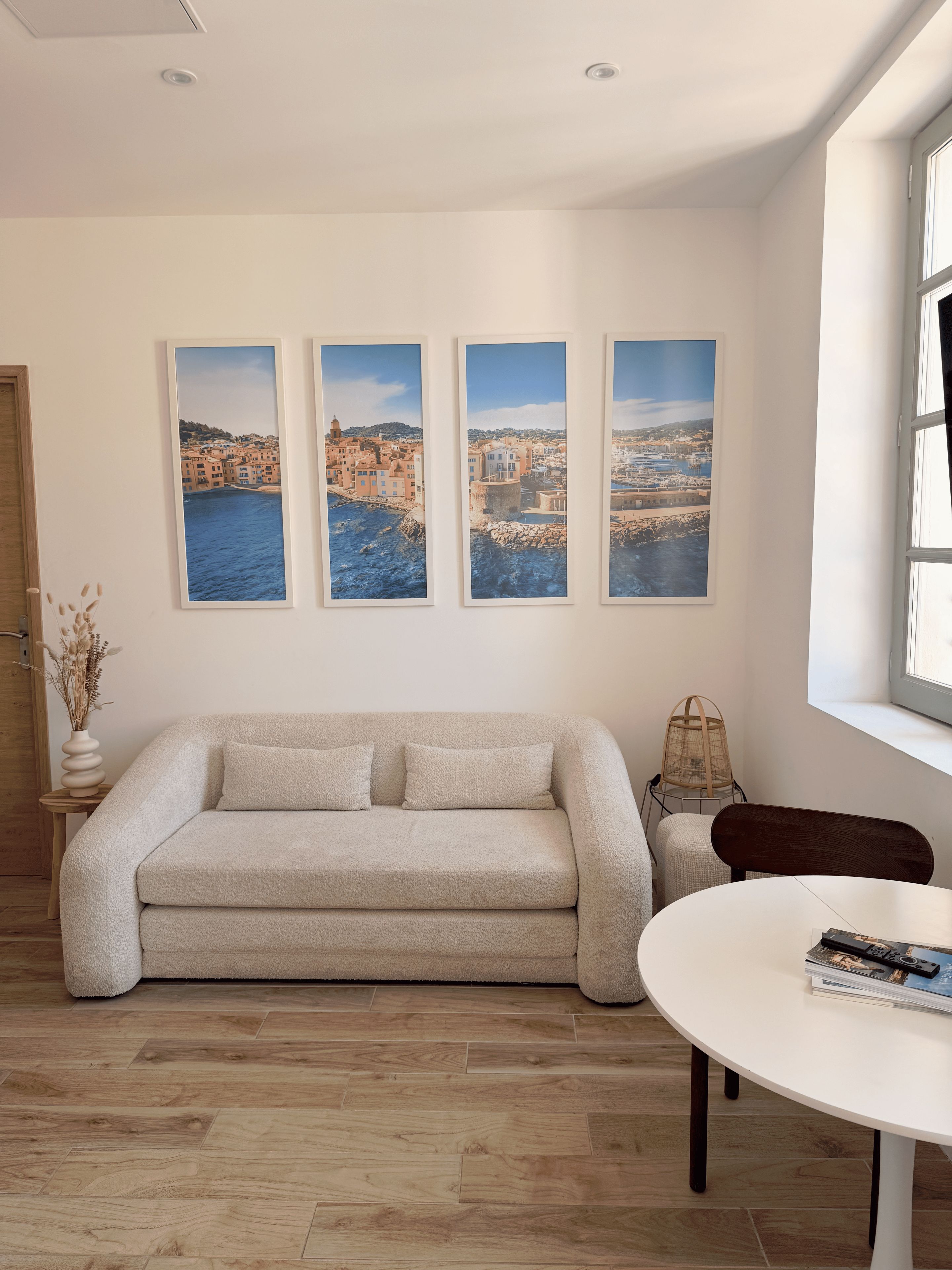 Una sala de estar con un sofá color beige, una mesa redonda blanca y tres fotografías enmarcadas de paisajes costeros en la pared.