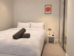 Een keurig opgemaakt bed met een opgerolde handdoek, een nachtkastje met een lamp en een ingelijst foto aan de muur.