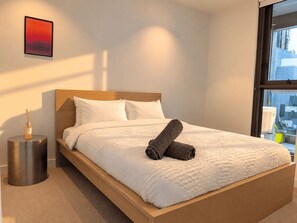 A neatly made bed with white bedding and a rolled towel, a small bedside table with a bottle, and a framed picture on the wall. - Future Past CBD Views & Balcony (Melbourne)