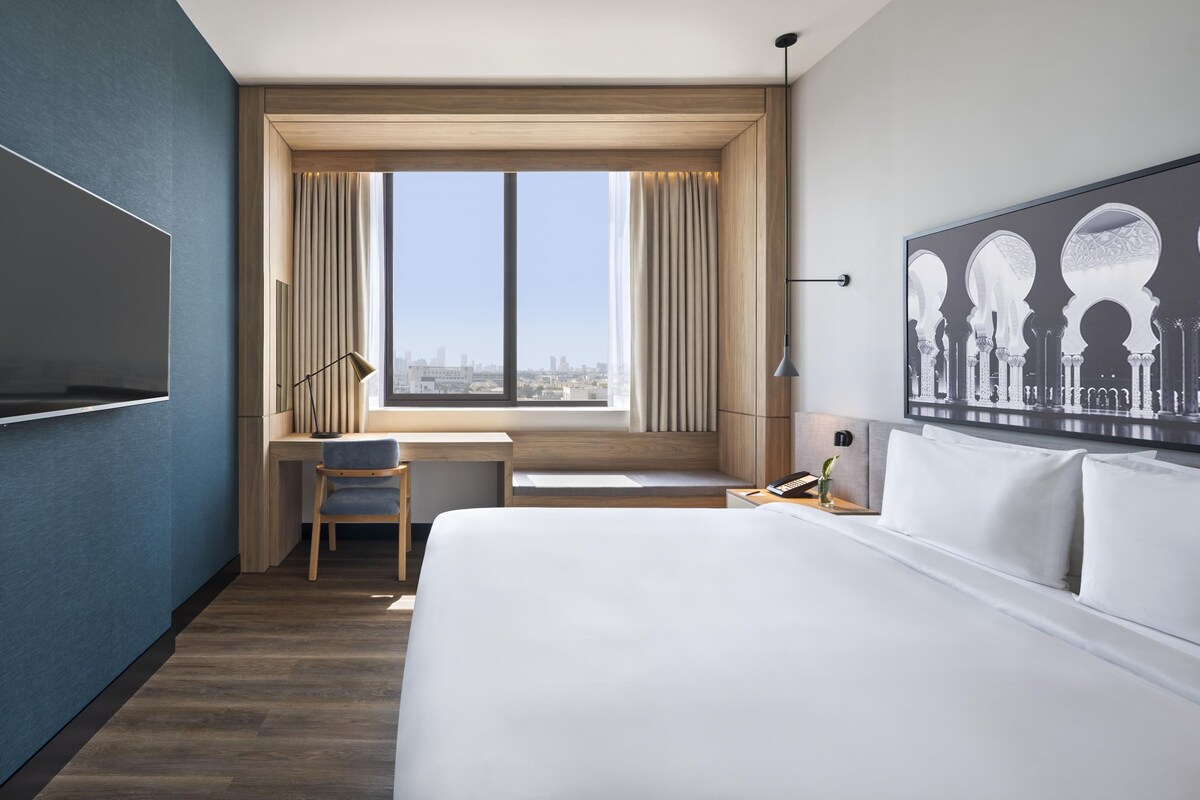A hotel room with a large bed, a desk, a chair, a TV, and a window with curtains.