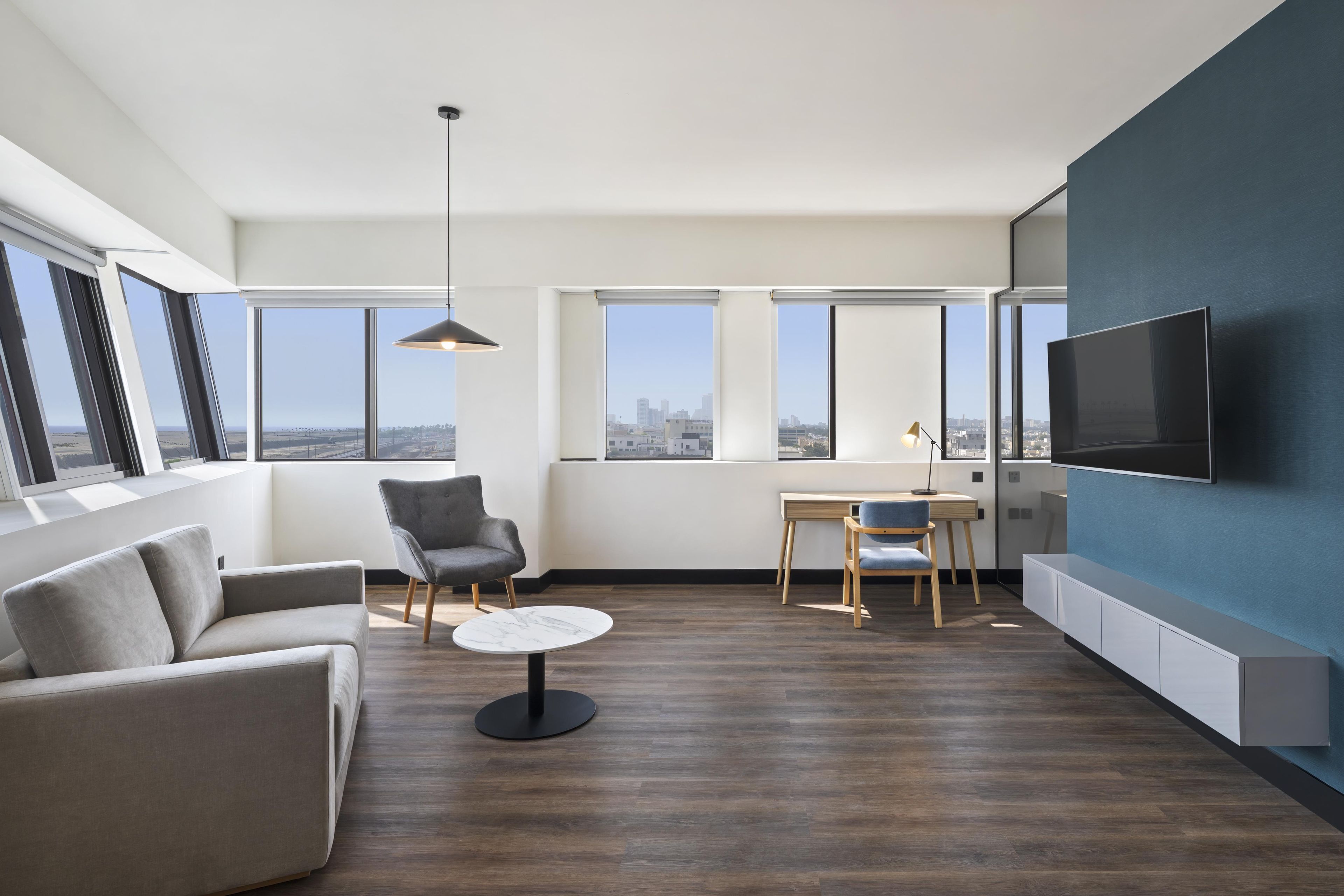 A modern living room with a sofa, a chair, a small table, a TV, and large windows offering a city view.