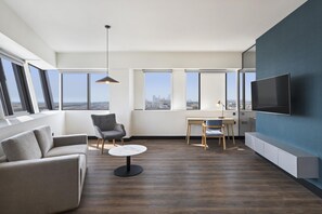 A modern living room with a sofa, a chair, a small table, a TV, and large windows offering a city view.