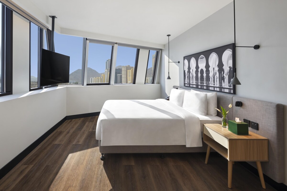 A modern hotel room with a large bed, a nightstand, a flat-screen TV, and a city view.