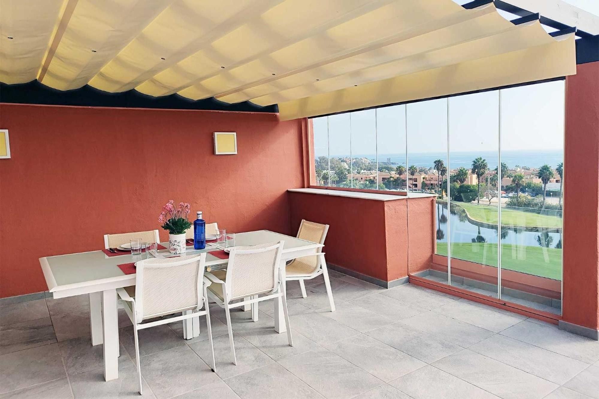 Uma área de jantar com mesa e cadeiras, uma janela ampla com vista para um campo de golfe e edifícios, e um teto com design diferenciado.