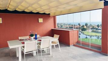 Uma área de jantar com mesa e cadeiras, uma janela ampla com vista para um campo de golfe e edifícios, e um teto com design diferenciado.