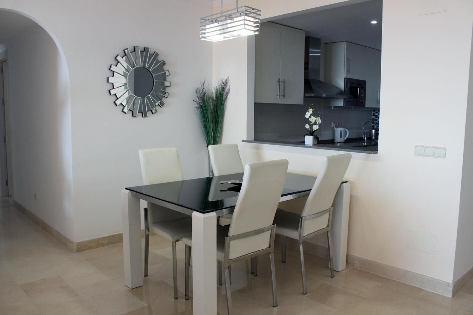Een moderne eetruimte met een glazen tafel en witte stoelen, een decoratieve spiegel en een keuken zichtbaar door een boog.
