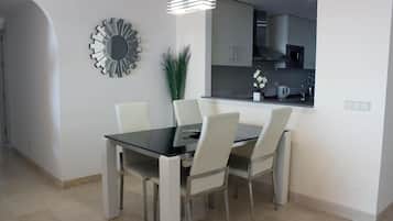 Een moderne eetruimte met een glazen tafel en witte stoelen, een decoratieve spiegel en een keuken zichtbaar door een boog.