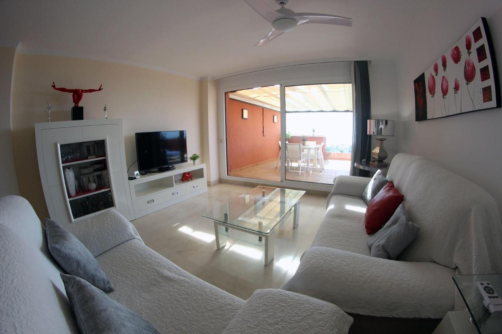 A living room with a sofa, a glass coffee table, a TV on a cabinet, and a balcony with a view.