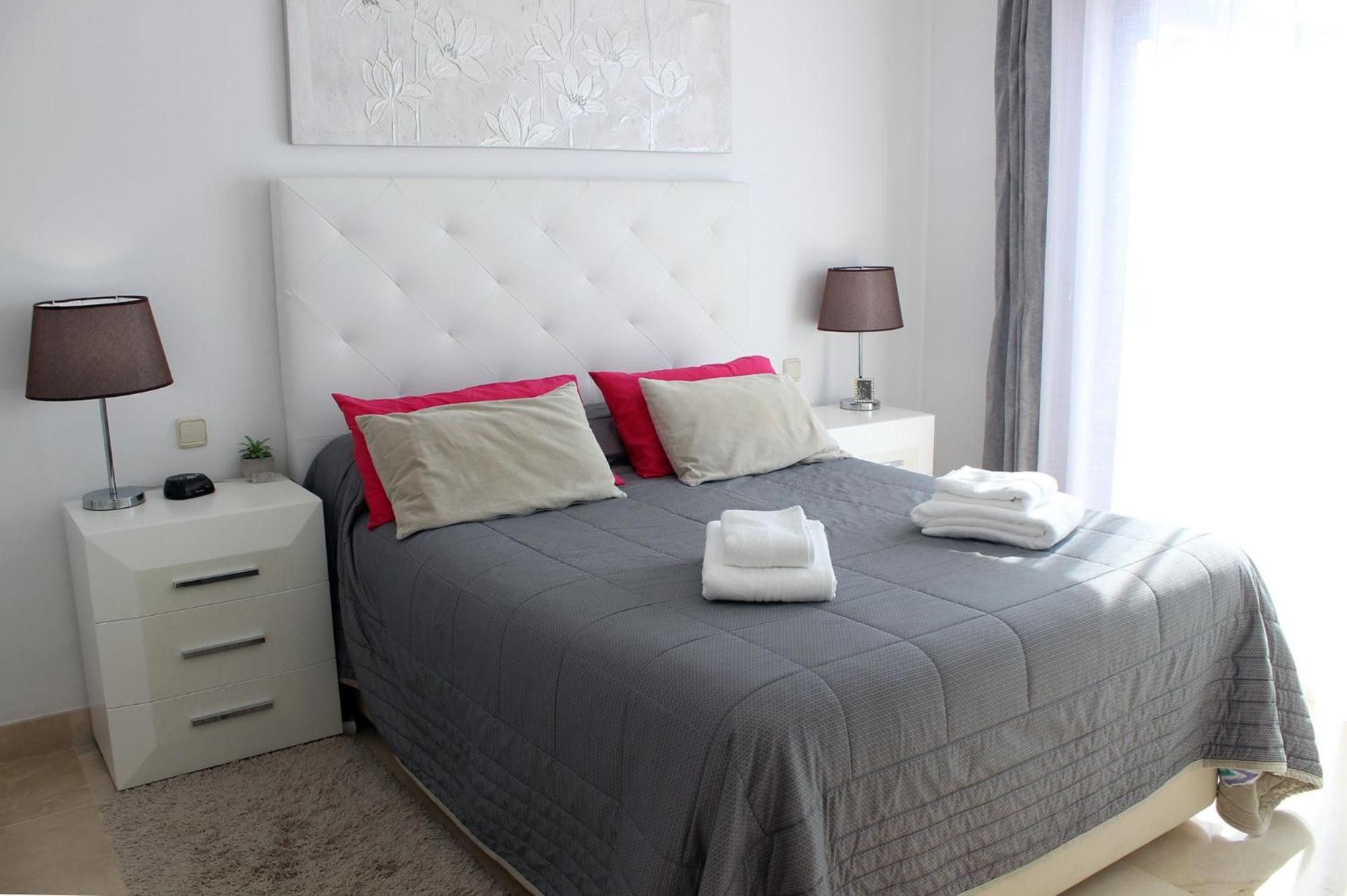 A neatly arranged hotel room with a bed, two bedside tables, a nightstand, a lamp, a wall art piece, and a window with curtains.