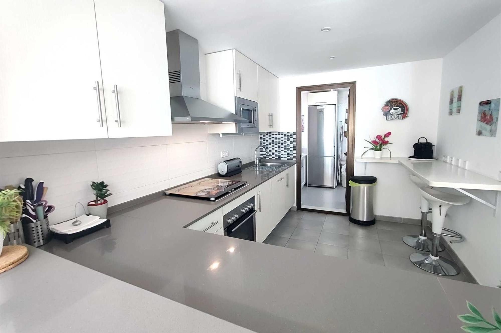Een moderne keuken met witte kasten, een roestvrijstalen koelkast en een klein eetgedeelte met een wit aanrecht en barstoelen.