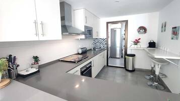 Een moderne keuken met witte kasten, een roestvrijstalen koelkast en een klein eetgedeelte met een wit aanrecht en barstoelen.