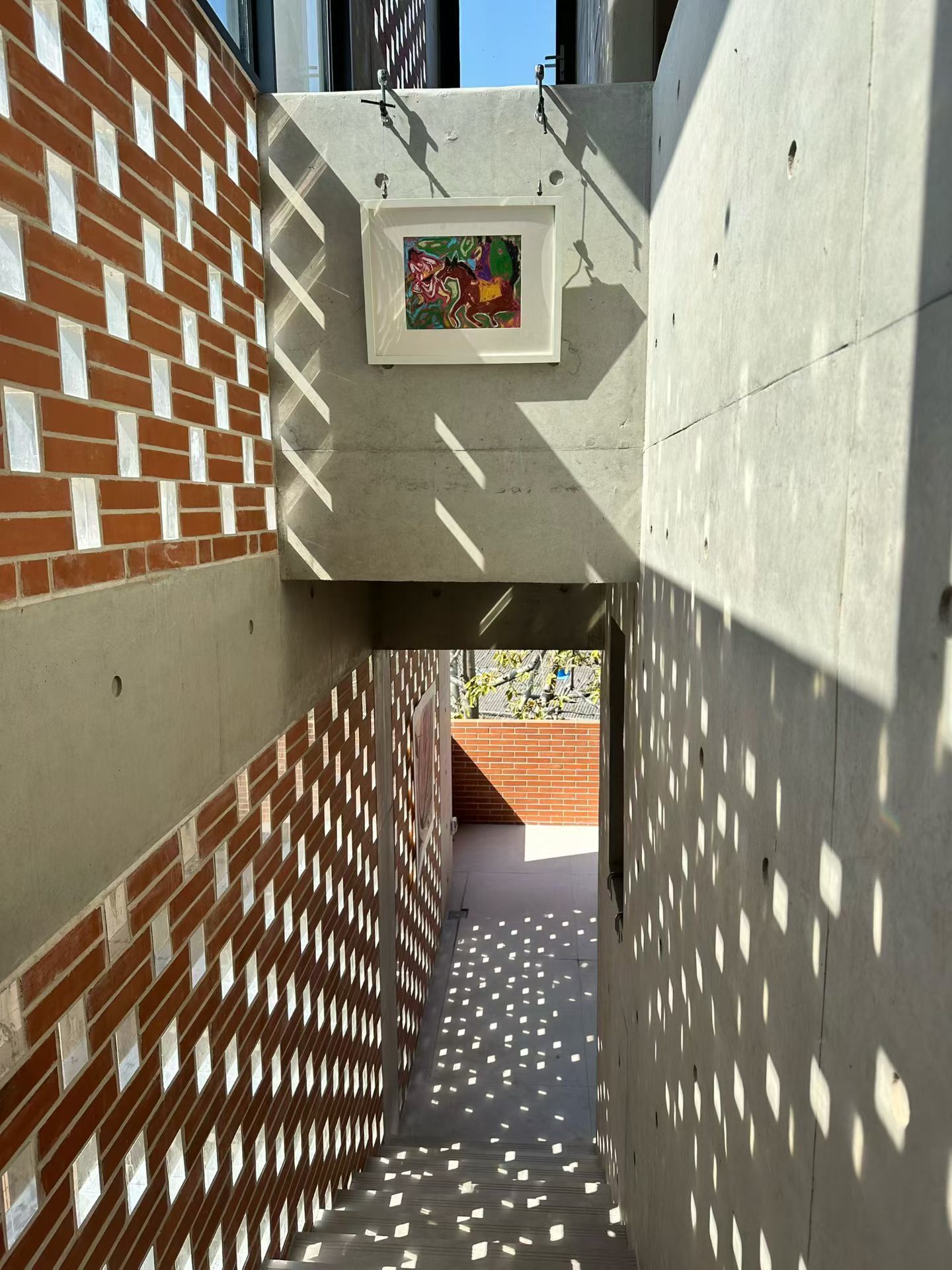 A narrow corridor with a brick wall on one side and a concrete wall on the other, leading to a door with a framed artwork.