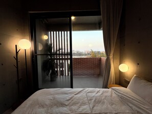 A bedroom with a bed, a potted plant, a balcony with a view, and two wall-mounted lamps.