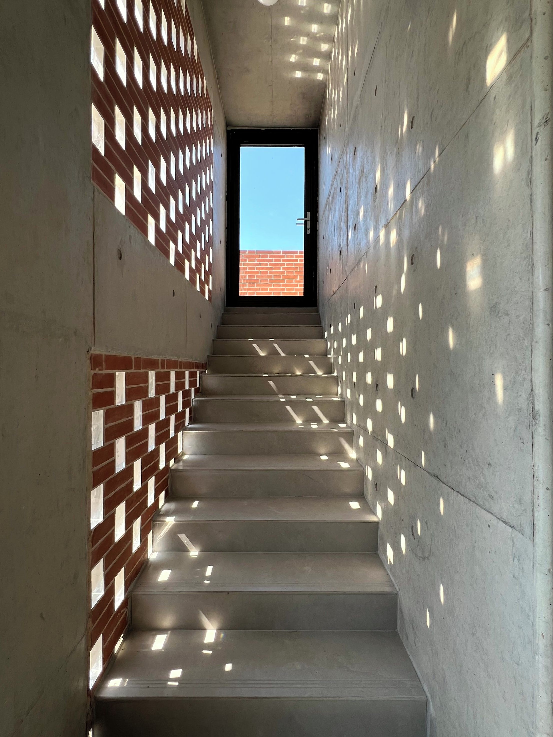 Un escalier aux murs en béton et aux éléments décoratifs en briques, menant à une porte qui offre une vue dégagée sur un bâtiment en briques situé à l’extérieur.