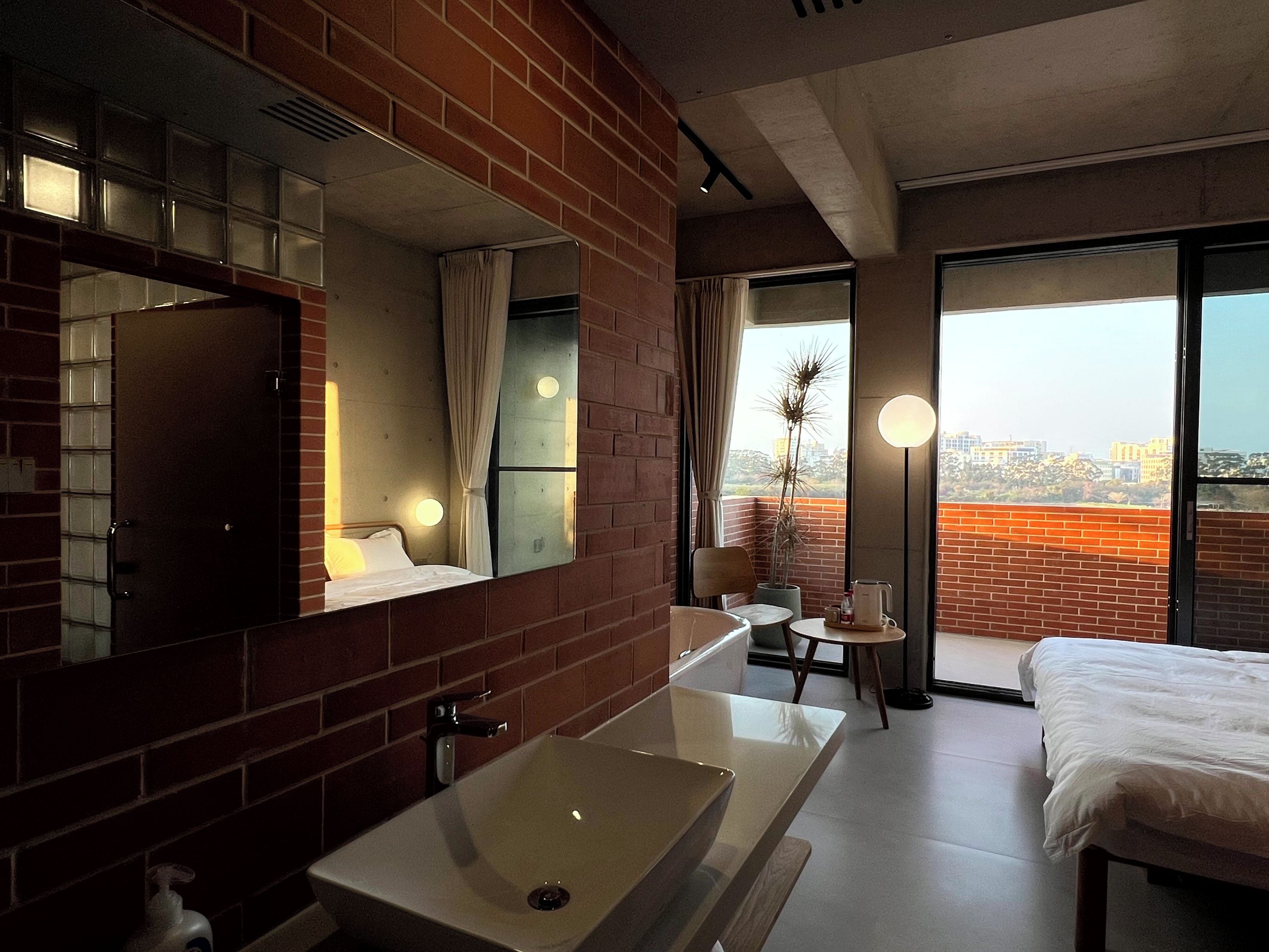 A hotel room with a large mirror, a white sink, a bed, a small table, and a view of the city through large windows.