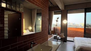 A hotel room with a large mirror, a white sink, a bed, a small table, and a view of the city through large windows.