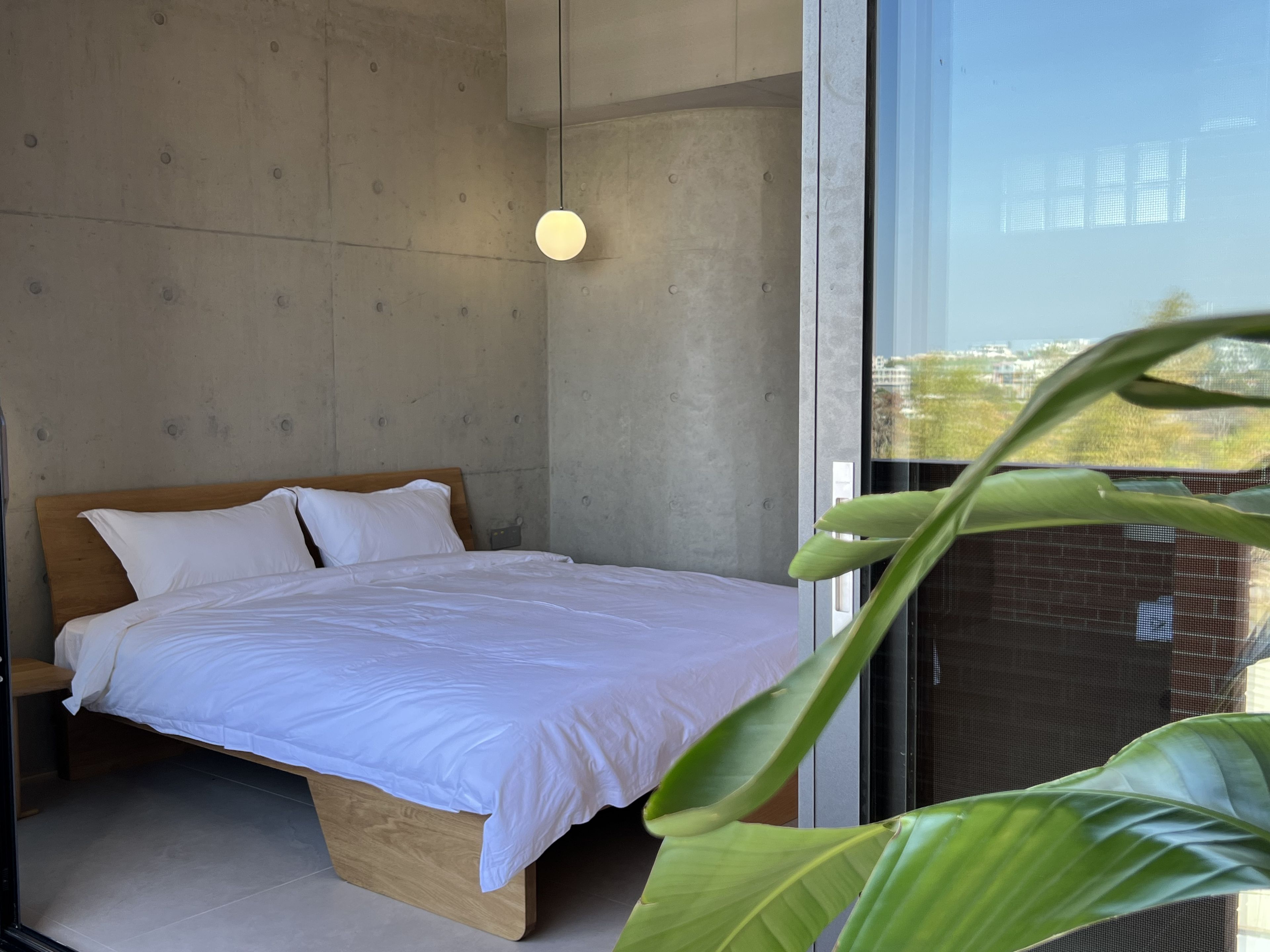 Un dormitorio moderno con cama de madera, ropa de cama blanca, una lámpara colgante y vistas por un gran ventanal.