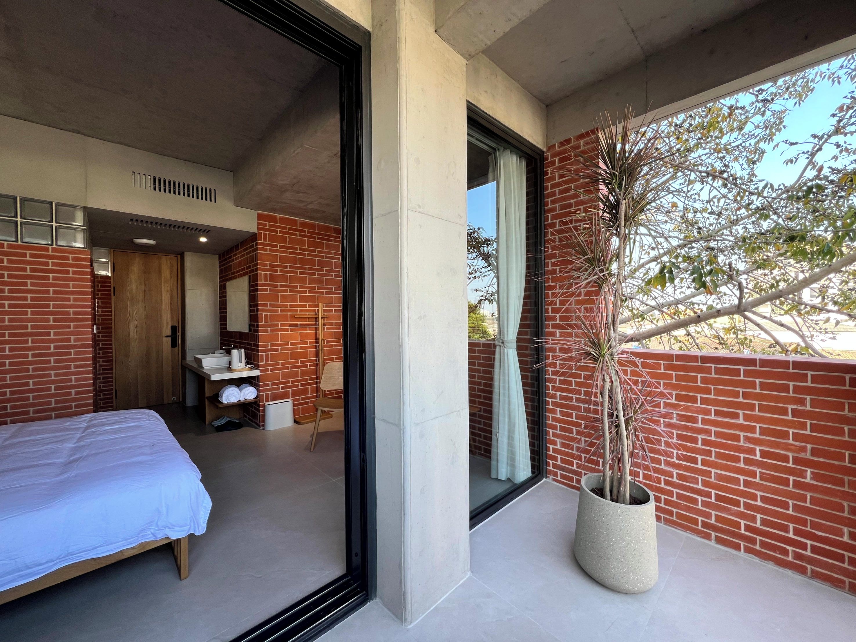 A modern bedroom with a large bed, a bathroom with a mirror and sink, and a balcony with a potted plant.