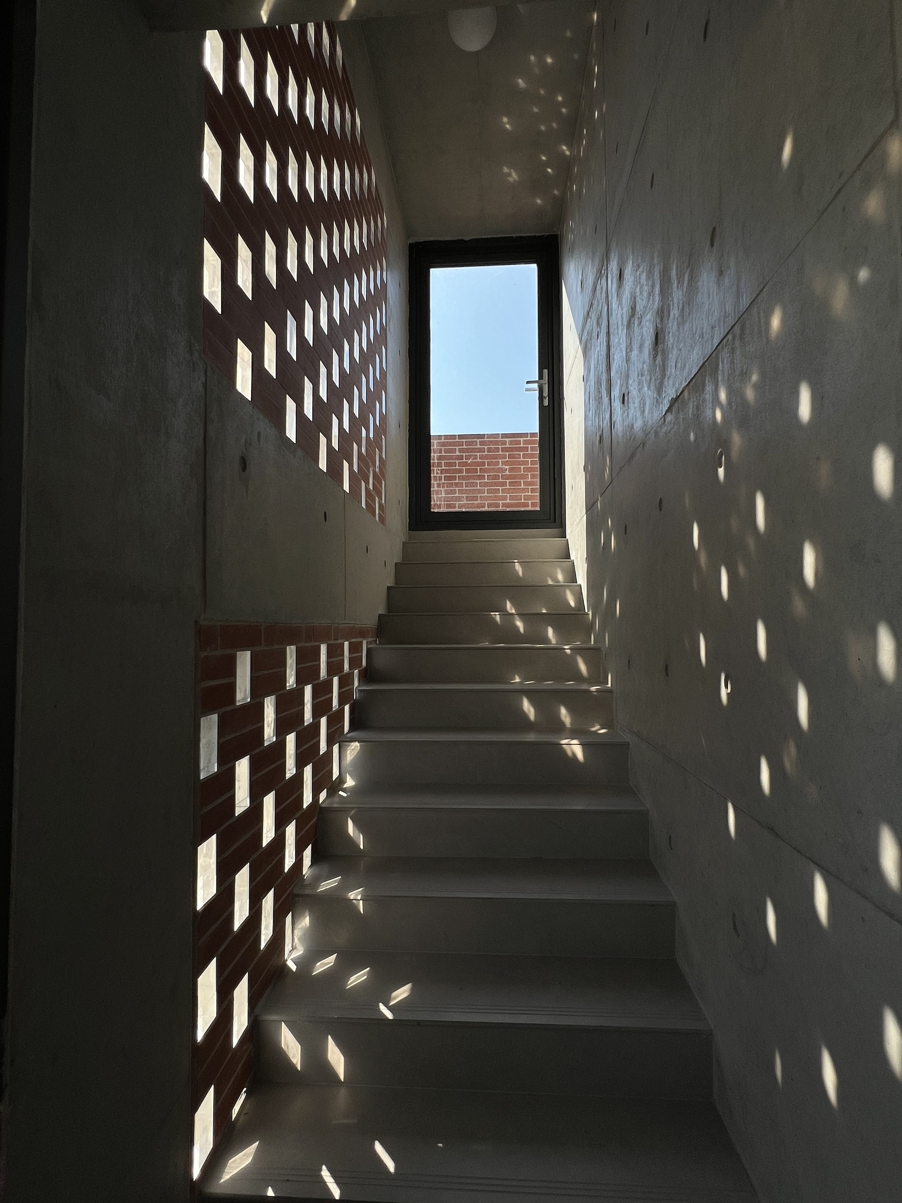 A staircase with a brick wall on one side and a concrete wall on the other, leading to a door with a view of a brick building outside.