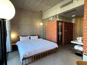 Habitación de hotel con cama, mesita de noche, lámpara, zona de ducha y una pequeña mesa con toallas.
