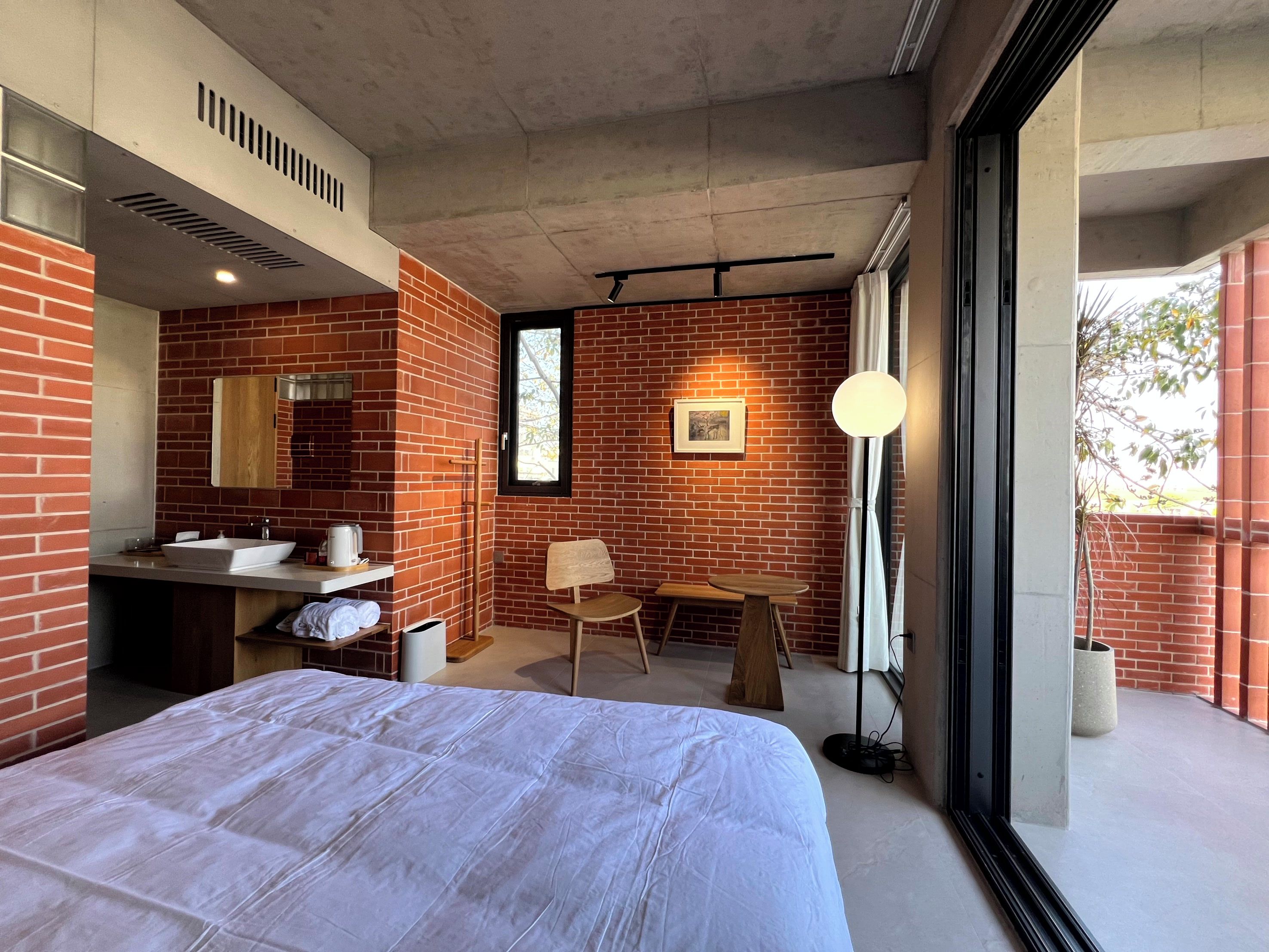A modern hotel room with a brick wall, a large bed, a vanity with a mirror, a small table, and a floor lamp.