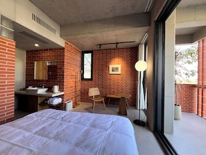 A modern hotel room with a brick wall, a large bed, a vanity with a mirror, a small table, and a floor lamp.