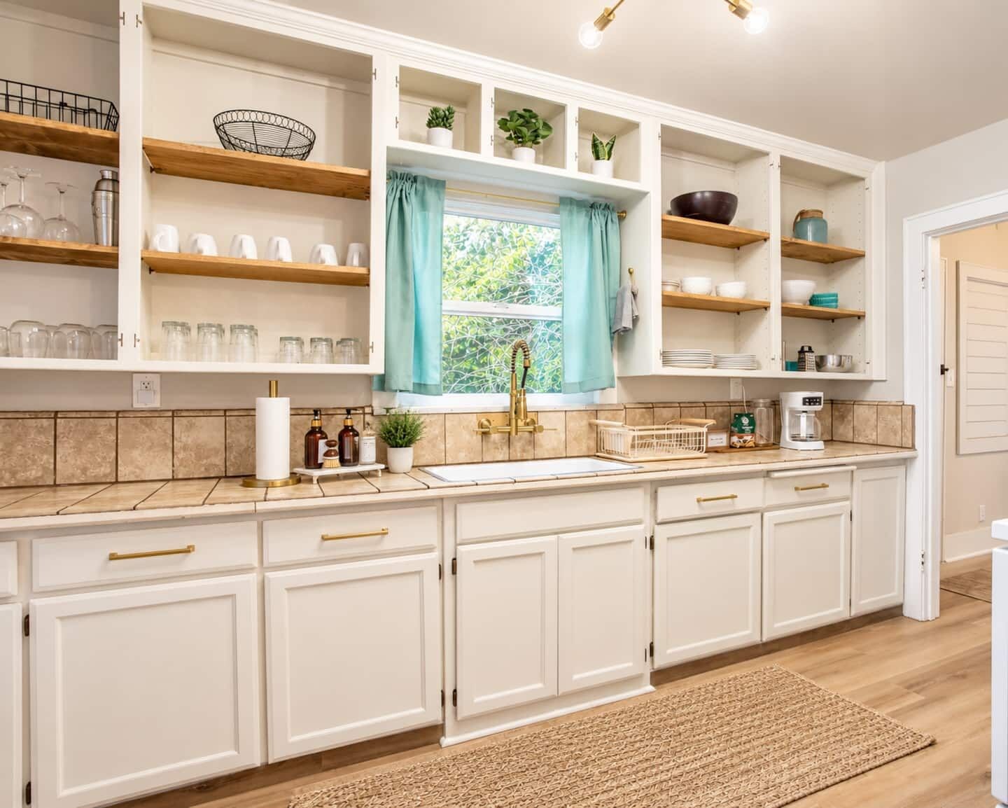A kitchen with white cabinetry, wooden shelves, a window with a curtain, and a kitchen island with a sink and coffee maker.
