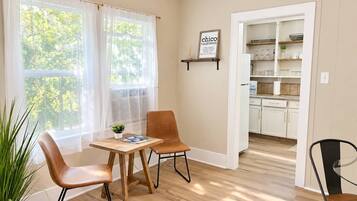A room with a dining table and chairs, a kitchen area visible through an open doorway, and a window with sheer curtains.