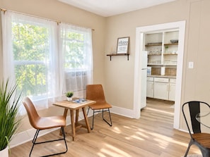 Ein Zimmer mit Esstisch und Stühlen, ein durchgehender Blick in eine Küchenzeile und ein Fenster mit durchscheinenden Vorhängen.
