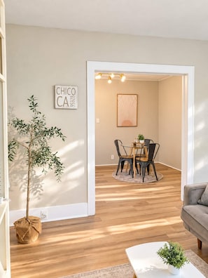 Una sala de estar con sofá, una mesita de centro y un comedor con mesa y sillas. Hay una planta en maceta y un cartel en la pared.