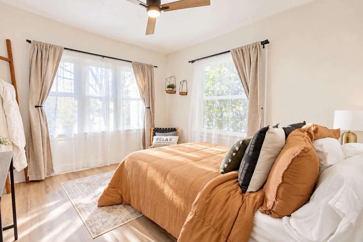 Un dormitorio con cama, cortinas, ventilador de techo, mesita de noche con lámpara y ventana con vista a árboles.