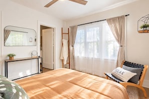 Un dormitorio con cama, una silla, un escritorio, un espejo y una ventana con cortinas.