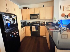 A compact kitchen with wooden cabinets, a black refrigerator, a microwave, and a sink. - Cozy 1-bedroom apartment in the heart of uptown Oakland with AC and gym access (Oakland)