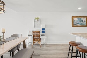 A dining area with a wooden table, two chairs, a wicker chair, a white cabinet, and a painting on the wall. - Renovated Direct Oceanfront Island Club (Hilton Head)