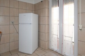 Un réfrigérateur blanc dans une pièce aux murs carrelés, avec une porte-fenêtre coulissante et un rideau à motifs floraux.