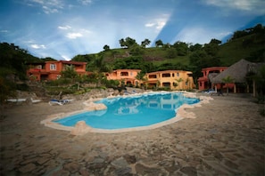 A pool surrounded by stone tiles, with multiple buildings in the background. - Case Damare Madrugada (las terrenas)