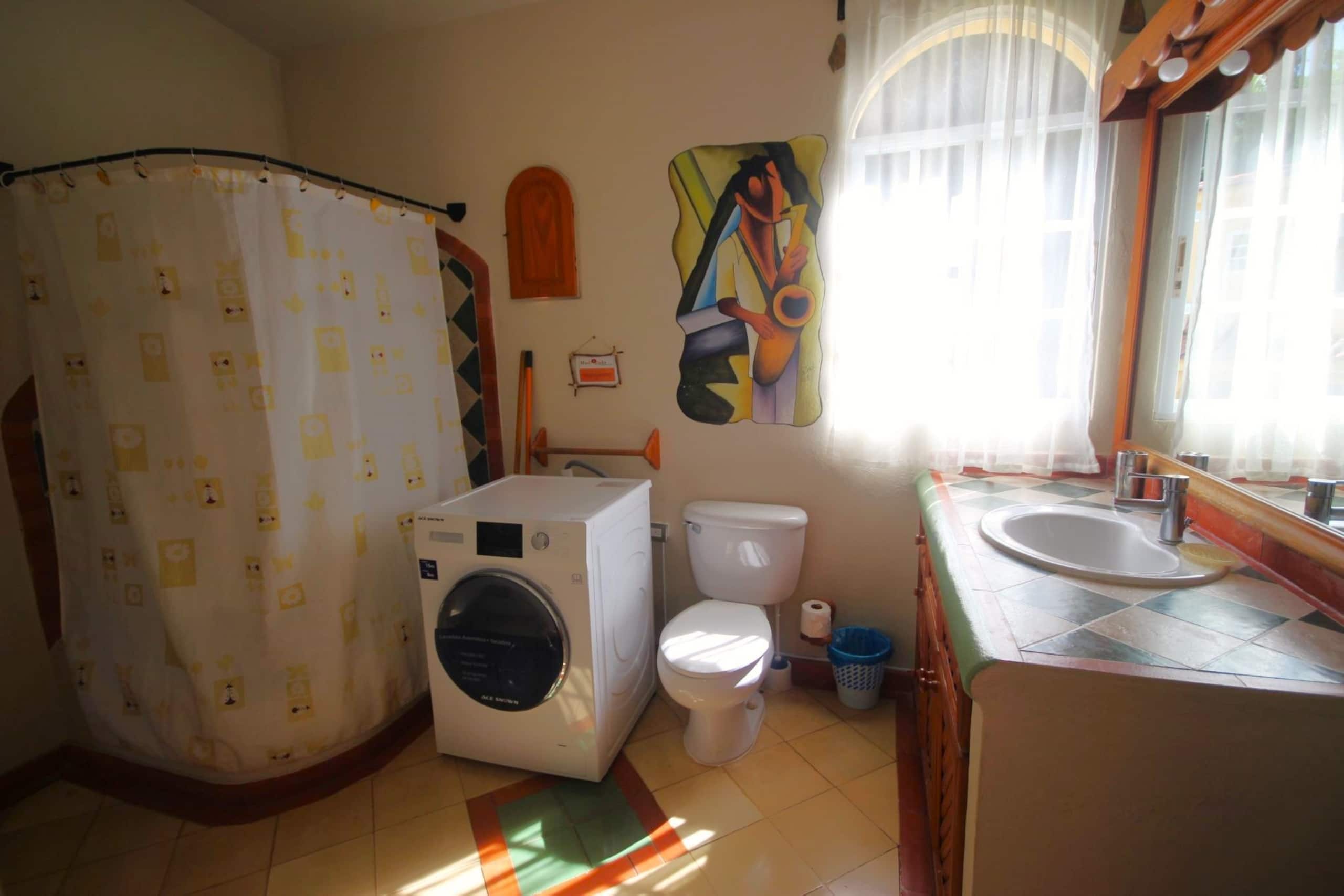 Un baño con lavadora, inodoro, cortina de ducha y un espejo grande.