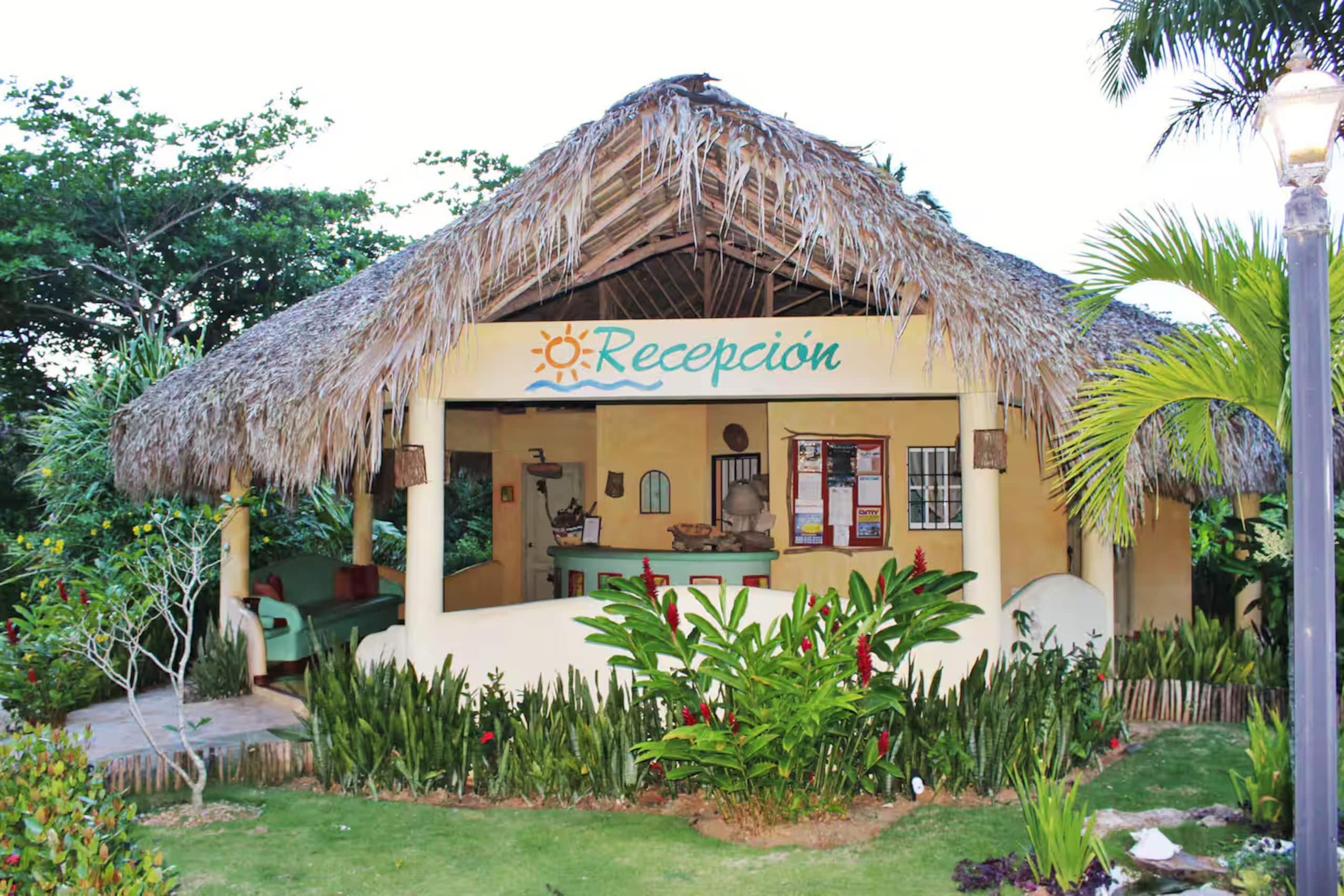 A thatched-roof reception building with a sign reading 'Recepcion' surrounded by tropical vegetation.