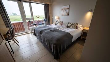 Ein Schlafzimmer mit einem großen Bett, einem Schreibtisch und einem Sessel. Durch das Fenster ist ein Garten zu sehen.